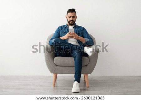 Young Arab guy using mobile phone, chatting online, sitting in armchair, surfing social media against white studio wall