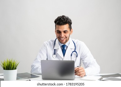 Young Arab Internist Working At Computer. Smiling Physician In White Coat With Stethoscope Sitting At Desk With Laptop. Professional Medical Consultation And Treatment In Hospital. Medical Healthcare.