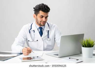 Young Arab Internist Working At Computer. Smiling Physician In White Coat With Stethoscope Sitting At Desk With Laptop. Professional Medical Consultation And Treatment In Hospital. Medical Healthcare.