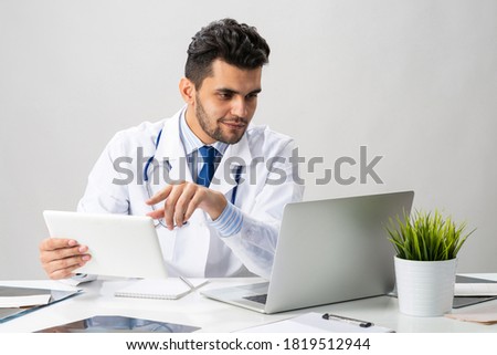 Young arab internist working with tablet computer. Physician in white medical gown with stethoscope sitting at desk with laptop. Professional medical consultation and treatment in modern clinic.