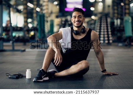 Young Arab Male Athlete Relaxing On Floor After Training At Gym, Smiling Middle Eastern Man Resting Next To Blank Container With Supplement Pills, Enjoying Bodybuilding And Healthy Lifestyle