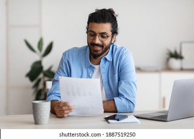 Young Arab Male Self-Entrepreneur Working With Papers At Desk In Home Office, Checking Annual Financial Reports. Eastern Millennial Man Reading Documents, Sitting At Table With Computer, Copy Space