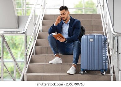 Young Arab Male Traveller Talking On Cellphone And Holding Passport, Rebooking Plane Tickets Online While Sitting With Luggage On Stairs In Airport Terminal, Middle Eastern Man Missed His Flight