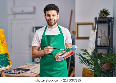 Young arab man artist smiling confident holding paintbrush   palette at art studio