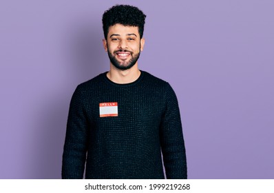 Young arab man with beard wearing hello my name is sticker identification looking positive and happy standing and smiling with a confident smile showing teeth 