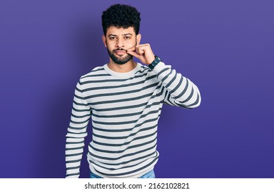 Young arab man with beard wearing casual striped sweater mouth and lips shut as zip with fingers. secret and silent, taboo talking 