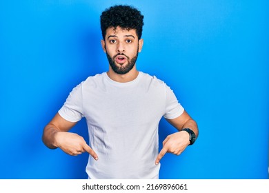 Young Arab Man With Beard Wearing Casual White T Shirt Pointing Down With Fingers Showing Advertisement, Surprised Face And Open Mouth 