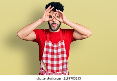 Young Arab Man With Beard Wearing Cook Apron Doing Ok Gesture Like Binoculars Sticking Tongue Out, Eyes Looking Through Fingers. Crazy Expression. 