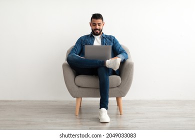 Young Arab Man In Casual Wear Working Online, Sitting In Armchair And Using Laptop Against White Studio Wall, Full Length. Millennial Eastern Guy Surfing Internet On Portable Pc