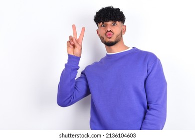 young arab man with curly hair wearing purple sweatshirt over white background makes peace gesture keeps lips folded shows v sign. Body language concept