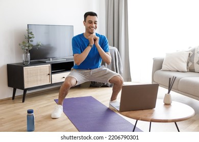 Young arab man doing deep squats, looking at laptop during online training, exercising at home, free space. Active guy working out in front of computer