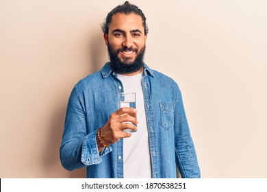 Young arab man drinking glass of water looking positive and happy standing and smiling with a confident smile showing teeth 