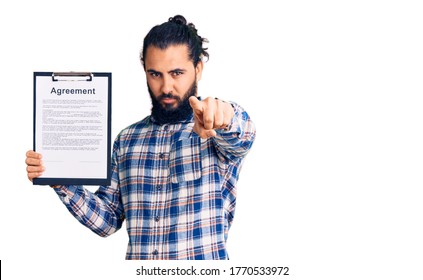 Young Arab Man Holding Clipboard With Agreement Document Pointing With Finger To The Camera And To You, Confident Gesture Looking Serious 