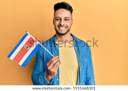 Young arab man holding costa rica flag looking positive and happy standing and smiling with a confident smile showing teeth 