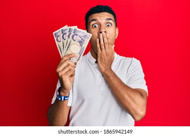 Young arab man holding japanese yen banknotes covering mouth with hand, shocked and afraid for mistake. surprised expression 