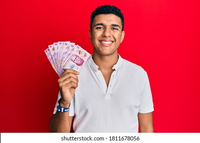 Young arab man holding mexican pesos looking positive and happy standing and smiling with a confident smile showing teeth 
