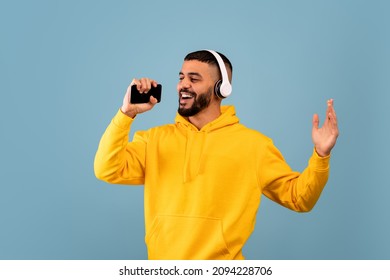 Young Arab Man Singing Holding Smartphone Like Microphone Wearing Wireless Headphones And Having Fun Listening To Music On Blue Studio Background. Online Karaoke Application Concept