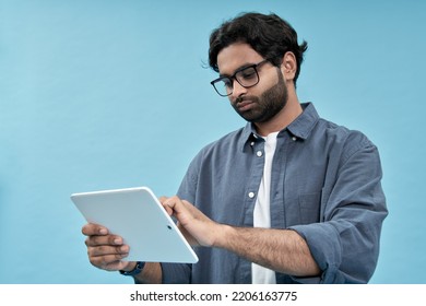 Young Arab Man Student Using Digital Tablet Elearning Standing Isolated On Blue Background. Ethnic Indian Guy Holding Pad Computer Wearing Glasses Watching Virtual Class Webinar, Browsing Internet.