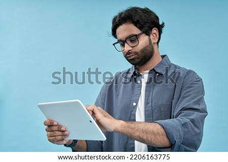 Young arab man student using digital tablet elearning standing isolated on blue background. Ethnic indian guy holding pad computer wearing glasses watching virtual class webinar, browsing internet.