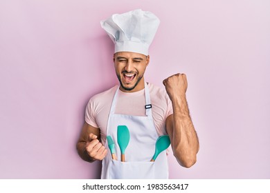 Joven árabe con delantal profesional de la cocina y sombrero que celebra sorprendido y asombrado por el éxito con los brazos levantados y los ojos cerrados 