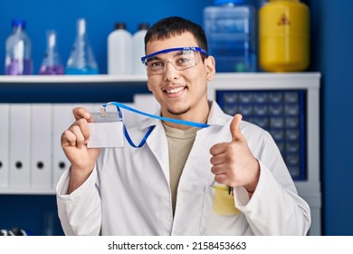 Young Arab Man Working At Scientist Laboratory Holding Id Card Smiling Happy And Positive, Thumb Up Doing Excellent And Approval Sign 