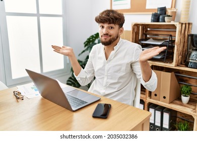 Young Arab Man Working Using Computer Laptop At The Office Clueless And Confused Expression With Arms And Hands Raised. Doubt Concept. 