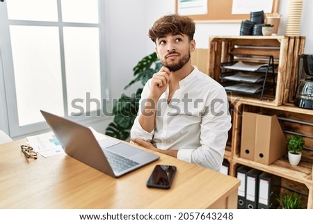 Young arab man working using computer laptop at the office serious face thinking about question with hand on chin, thoughtful about confusing idea 