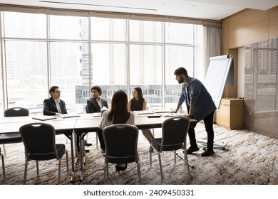 Young Arab project manager man presenting startup to colleagues, standing at large meeting table at leader place. Coach giving seminar, lecture in modern office boardroom interior