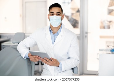 Young Arab Stomatologist Doctor Using Digital Tablet At Workplace In Clinic, Middle Eastern Dentist Wearing Protective Medical Mask And Uniform Posing With Tab Computer In Stomatological Cabinet