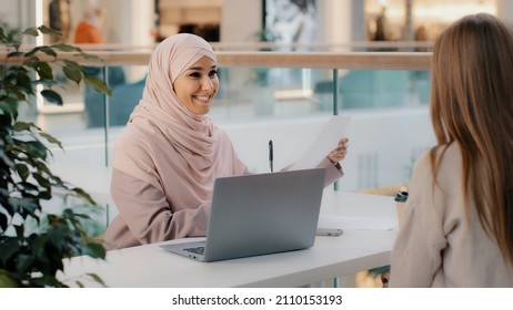 Young arab woman consultant sales agent bank worker sitting at office desk advises unrecognizable girl client offers to sign contract sale agreement helps to arrange loan proposes legal consultation