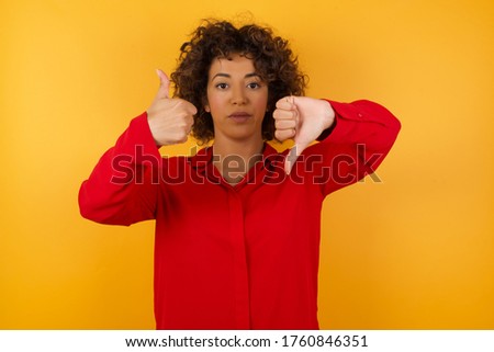 Young arab woman with curly hair wearing red shirt  over yellow background showing thumbs up and thumbs down, difficult choose concept 
