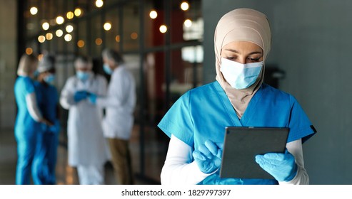 Young Arab Woman Doctor In Hijab, Medical Mask And Gloves Standing In Hospital And Using Tablet Device. Muslim Female Medic In Traditional Headscarf Tapping On Gadget Computer In Clinic. Covid-19.