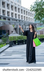 Young Arab Woman Shopping Outdoors Wearing Abaya Pointing And Carrying Shopping Bags In The City