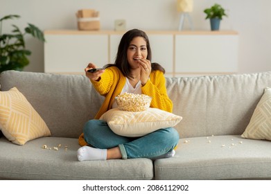 Young arab woman sitting on couch with TV remote, choosing movie to watch and eating popcorn, enjoying free time, copy space. Positive middle eastern lady switching channels