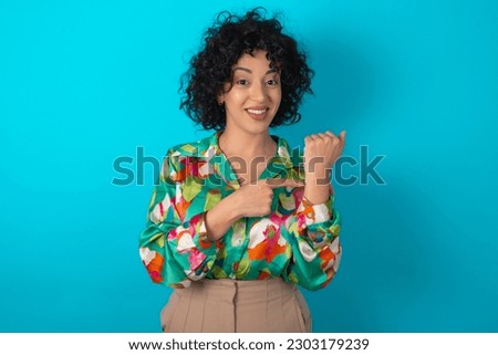 young arab woman wearing colorful shirt over blue background In hurry pointing to wrist watch, impatience, looking at the camera with relaxed expression