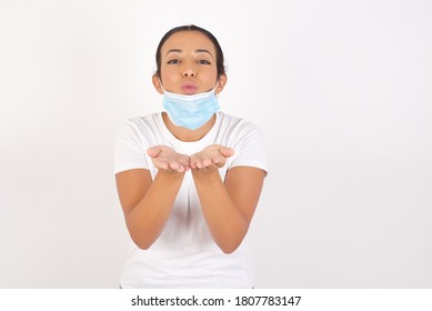 Young Arab Woman Wearing Medical Mask Standing Over Isolated White Background Sending Blow Kiss With Pout Lips And Holding Palms To Send Air Kiss.