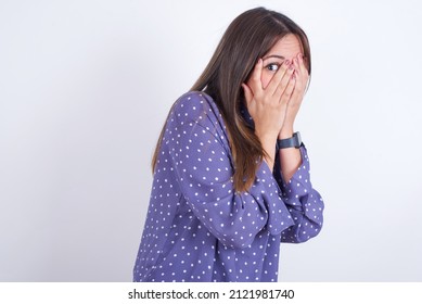 Young arab woman wearing polka-dot clothes over white backgtound covering face with hands and peering out with one eye between fingers. Scared from something or someone.