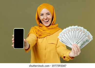 Young arabian asian muslim woman in abaya hijab yellow clothes hold mobile phone fan cash money dollar banknotes screen workspace isolated on olive green background People uae islam religious concept