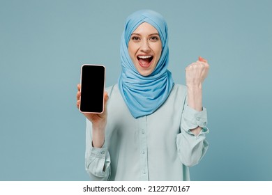 Young arabian asian muslim woman in abaya hijab hold in hand use mobile cell phone blank screen area do winner gesture isolated on plain blue background. People middle eastern islam religious concept