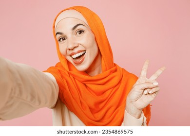 Young arabian asian muslim woman wears orange abaya hijab doing selfie shot pov on mobile cell phone isolated on plain light pink background studio portrait. Uae middle eastern islam religious concept - Powered by Shutterstock