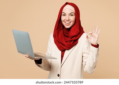 Young Arabian Asian Muslim IT woman wear red abaya hijab suit clothes hold use work on laptop pc computer show ok okay isolated on plain beige background. UAE middle eastern Islam religious concept - Powered by Shutterstock