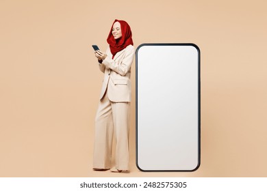 Young Arabian Asian Muslim woman wear red abaya hijab suit clothes stand big huge blank screen free area mobile cell phone use smartphone device isolated on plain beige background. UAE Islam concept - Powered by Shutterstock