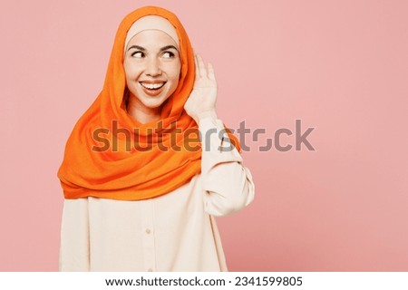 Young arabian asian muslim woman wear orange abaya hijab try to hear you overhear listening intently isolated on plain light pink background studio. People uae middle eastern islam religious concept