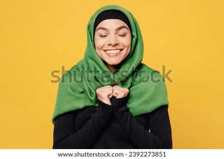 Young arabian asian muslim woman wear green hijab abaya black clothes hold hands folded clench fist close eyes isolated on plain yellow background. People uae middle eastern islam religious concept