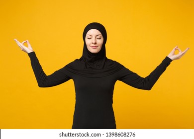 Young arabian muslim woman in hijab black clothes hold hands in yoga gesture, relax meditating isolated on yellow background, studio portrait. People religious lifestyle concept. Mock up copy space