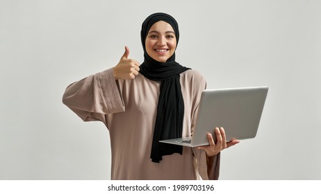 Young arabian woman in hijab showing thumb up and smiling at camera while posing with laptop on light background, widescreen. Beautiful muslim lady