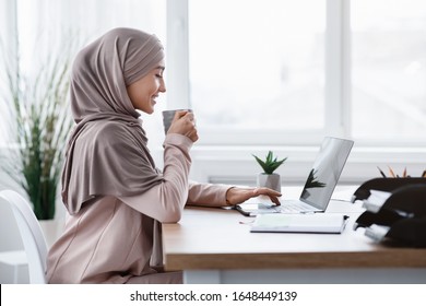 Young arabic businesswoman enjoying cup of coffee at her desk while working on laptop in modern office, side view with free space