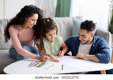 Young Arabic Parents And Their Cute Little Daughter Drawing Together At Home. Middle Eastern Mom And Dad Painting With Their Preschool Child In Living Room, Using Pencils And Having Fun, Free Space