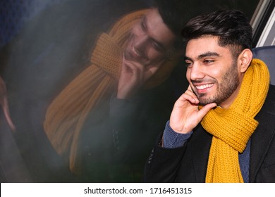 Young Arabic Smiling Positive Traveler Handsome Man Wearing Black Coat And Yellow Scarf Sitting On Chair And Looks Into Window In Fast Modern German Train At Night. Travel And Transportation Concept