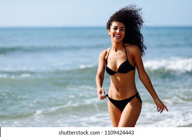Young arabic woman with beautiful body in swimwear smiling in a tropical beach. Brunette female with curly long hairstyle wearing black bikini.
